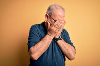 Orta yaşlı yakışıklı beyaz adam sarı arka plan üzerine gündelik polo ve gözlük takıyor ve ağlarken suratını ellerle kapatıyor. Depresyon kavramı.