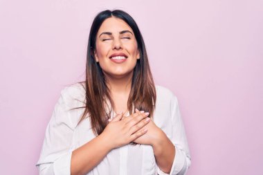 Zarif bir gömlek giyen güzel esmer kadın izole edilmiş pembe arka planda duruyor. Elleri göğsünde gülümsüyor. Yüzünde minnet dolu bir jest ile gözleri kapalı. Sağlık kavramı.