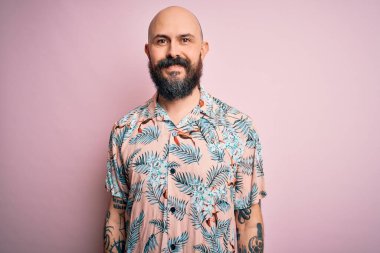 Sakallı ve dövmeli yakışıklı, pembe arka planda sıradan bir çiçek gömleği giyen, yüzünde mutlu ve havalı bir gülümseme olan kel bir adam. Şanslı kişi..