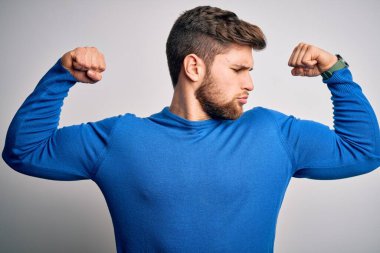 Sakallı, mavi gözlü, sıradan süveter giyen ve kollarında gururla gülümseyen genç sarışın adam. Fitness konsepti.