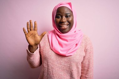Genç Afro-Amerikan artı cüsseli kadın tecrit edilmiş pembe arka plan üzerine Müslüman başörtüsü takıyor ve beş numarayı gösterirken kendinden emin ve mutlu gülümsüyor..