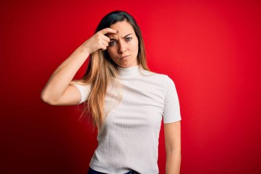 Kırmızı arkaplan üzerine sıradan beyaz tişörtler giyen güzel sarışın kadın alnındaki sivilceden mutsuz olduğunu gösteriyor, siyah noktalı çirkin bir enfeksiyon. Sivilce ve deri problemi