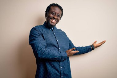 Genç, yakışıklı, Afro-Amerikan bir adam, günlük gömleğini giymiş beyaz arka planda duruyor ve gülümseyerek içeri girmeye davet ediyor.