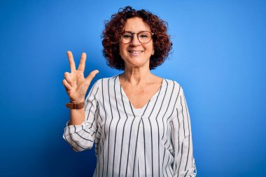 Orta yaşlı, kıvırcık saçlı, rahat çizgili tişört giyen, izole edilmiş arka planda üç numaralı parmaklarıyla gülümseyerek kendinden emin ve mutlu bir şekilde duran güzel bir kadın..