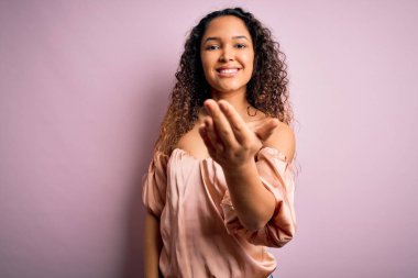 Kıvırcık saçlı, rahat tişörtlü, pembe arka planda duran genç ve güzel bir bayan selamlaşma ve dostça el sıkışma sunuyor. Başarılı bir iş..