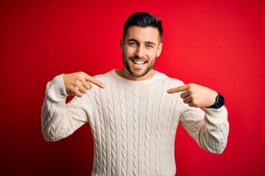 İzole edilmiş kırmızı arka plan üzerinde duran, rahat beyaz süveter giyen genç yakışıklı bir adam yüzünde bir gülümsemeyle gururlu ve mutlu bir şekilde kendini işaret ediyor..