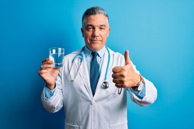 Orta yaşlı, yakışıklı, gri saçlı doktor. Mavi arka planda bir bardak su içiyor. Kocaman gülümsüyor.