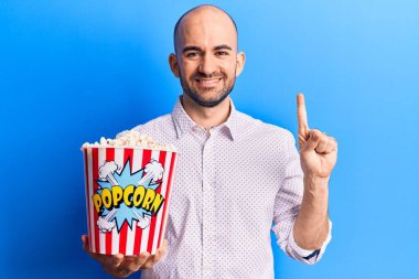 Genç ve kel bir adam elinde patlamış mısır tutarken bir fikir ya da soruyla gülümsüyor. 