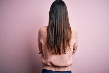 Genç, güzel, esmer kadın, pembe arka plan üzerine günlük süveter giyip kollarını kavuşturup arkana bakıyor.