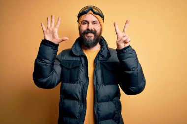 Sakallı, kar maskesi takmış, kar maskesi takmış, kel yakışıklı kayakçı yedi numaralı parmaklarıyla gösteriş yapıyor. Kendinden emin ve mutlu gülümsüyor..