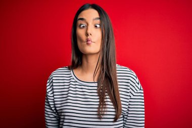 Genç, güzel, esmer bir kadın kırmızı arka planda günlük çizgili tişört giyiyor. Dudaklı balık suratı yapıyor, çılgınca ve komik bir jest. Komik ifade.