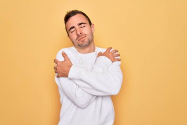 Mavi gözlü genç adam sarı arka planda dikilip kendine sarılıyor mutlu ve pozitif, kendinden emin gülümsüyor. Sevgi ve özgüven.