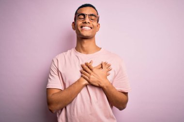 Yakışıklı Afro-Amerikalı adam pembe arka planda günlük tişört ve gözlük takıyor elleri göğsünde, gözleri kapalı ve yüzünde minnet dolu bir jest ile gülümsüyor. Sağlık kavramı.