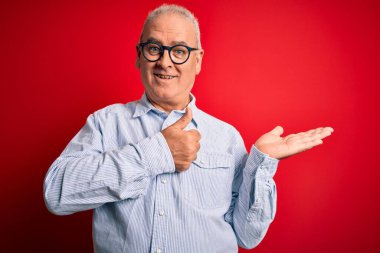 Orta yaşlı, beyaz, sıradan çizgili gömlek giyen, kırmızı arka planda gözlük takan, avucunu gösteren ve baş parmaklarıyla hareket eden mutlu ve neşeli gülümseyen yakışıklı bir adam.