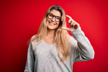 İzole edilmiş kırmızı arka plan üzerinde süveter ve gözlük takan genç ve güzel sarışın kadın gülümsüyor ve kendine güvenen el hareketleriyle küçük beden işareti yapıyor parmaklarıyla bakıyor ve kameraya bakıyor. Ölçü kavramı.