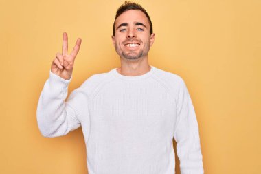 Mavi gözlü, sıradan bir süveter giyen, sarı arka planda duran, iki numaralı parmaklarıyla işaret eden, kendinden emin ve mutlu gülümseyen genç yakışıklı bir adam..