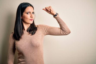 Young brunette woman with blue eyes wearing casual sweater over isolated white background Strong person showing arm muscle, confident and proud of power