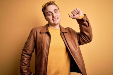 Genç, kızıl saçlı, günlük deri ceket giyen, izole edilmiş sarı arka planda gerinen, yorgun ve rahat, uykulu ve sabahın erken saatlerinde esneyen genç bir adam.