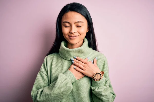 孤立したピンクの背景の上にタートルネックセーターを身に着けている若い美しい中国の女性は 目を閉じて胸に手で笑顔と顔に感謝のジェスチャー 健康コンセプト — ストック写真