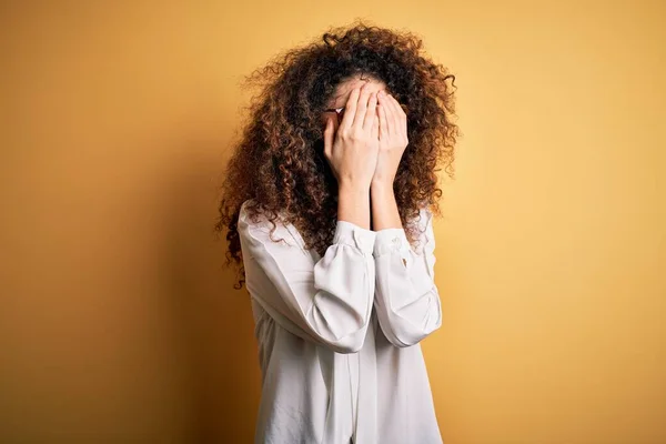 Young beautiful brunette woman with curly hair and piercing wearing shirt and glasses with sad expression covering face with hands while crying. Depression concept.