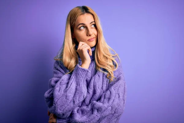 若い美しいブロンドの女性は 質問について考える顎の手で紫色の背景にカジュアルなタートルネックセーターを身に着けています 集中的な表情 思慮深い顔で微笑んだ 疑いの概念 — ストック写真