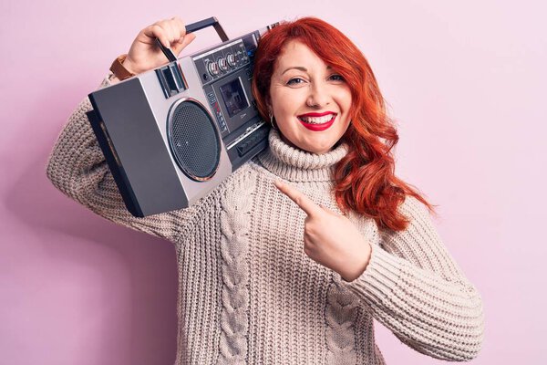 Beautiful redhead woman listening to music holding vintage boombox over pink background smiling happy pointing with hand and finger