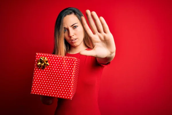 Young beautiful blonde woman with blue eyes holding birthday gift over red background with open hand doing stop sign with serious and confident expression, defense gesture