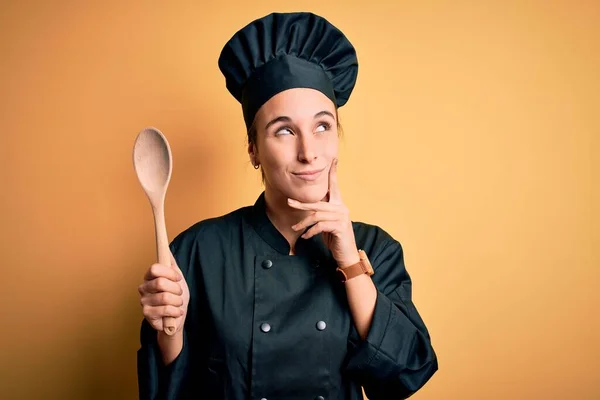 Young beautiful chef woman wearing cooker uniform and hat holding wooden spoon serious face thinking about question, very confused idea
