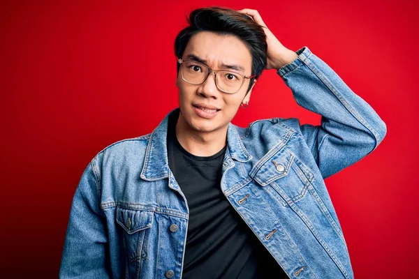 Young handsome chinese man wearing denim jacket and glasses over red background confuse and wonder about question. Uncertain with doubt, thinking with hand on head. Pensive concept.