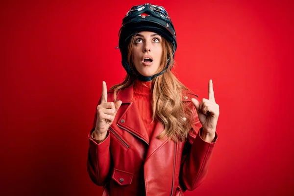 若いです美しいですブルネットMotoヘルメットを身に着けているオートバイの女性上の赤い背景驚きと驚きを見上げますと指と上げ腕 — ストック写真