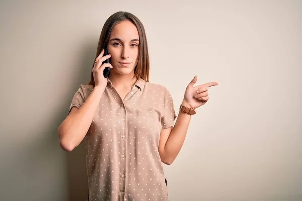 白い背景の上にスマートフォンで会話をしている若い美しい女性広告 深刻で穏やかな顔を示す側に手指で指して — ストック写真