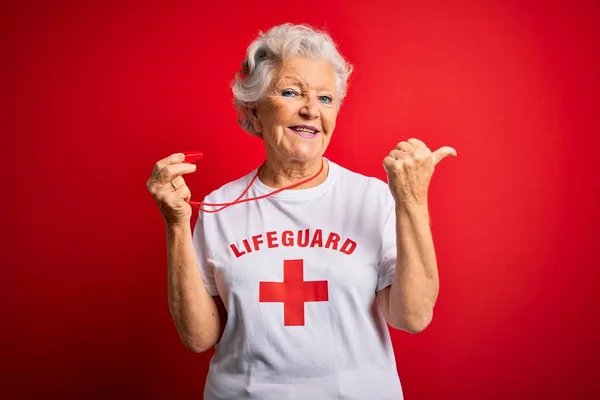 赤い十字のTシャツを着たシニアの美しい白髪のライフガードの女性が笛を使用して手と親指を後ろに向け 自信を持って笑顔 — ストック写真