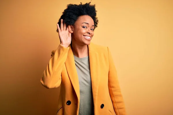 Young beautiful African American afro businesswoman with curly hair wearing yellow jacket smiling with hand over ear listening an hearing to rumor or gossip. Deafness concept.