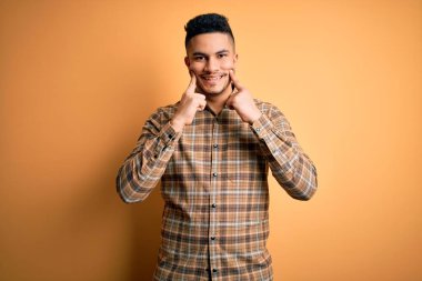 Sarı arka plan üzerinde duran, rahat gömlekli genç yakışıklı adam ağzı açık gülümsüyor, parmakları işaret ediyor ve neşeli bir gülümseme sergiliyor.