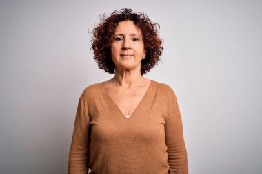 Orta yaşlı, kıvırcık saçlı güzel bir kadın izole edilmiş beyaz arka plan üzerine sıradan bir kazak giyiyor ve yüzünde ciddi bir ifade var. Basit ve doğal bir şekilde kameraya bakıyorsun..