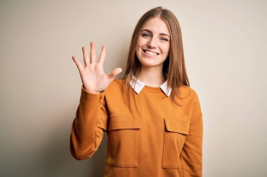 Genç, güzel, kızıl saçlı bir kadın izole edilmiş beyaz bir arka plan üzerine günlük süveter giyiyor ve beş numaralı parmaklarıyla işaret ederken kendinden emin ve mutlu gülümsüyor..