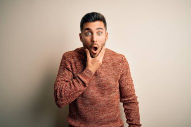 İzole edilmiş beyaz arka plan üzerinde duran sıradan bir süveter giyen genç yakışıklı elleri çenesinde olan inançsızlıktan, şaşkınlıktan ve şaşkınlıktan büyülenmiş görünüyor.