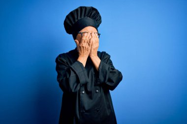 Kıdemli, gri saçlı, aşçı üniforması giyen, mavi arka planda şapka takan, ağlarken yüzünü ellerle kapatan güzel bir aşçı kadın. Depresyon kavramı.