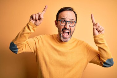 Orta yaşlı, ağarmış, günlük süveter ve gözlük takan, izole edilmiş sarı arka planda gülümseyen, şaşkın, parmaklarıyla işaret eden ve kollarını kaldıran bir adam..