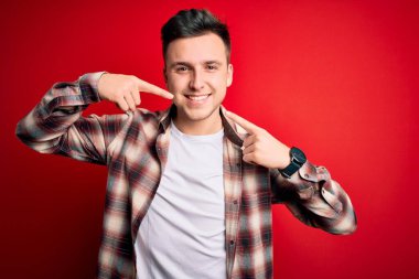 Kırmızı izole edilmiş arka plan üzerine gündelik modern tişört giyen, neşeli gülümseyerek parmaklarını ve ağzını gösteren genç beyaz bir adam. Diş sağlığı konsepti.