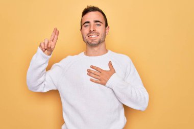 Mavi gözlü, sıradan bir süveter giyen, sarı arka planda durup gülümseyen, elleri göğsünde ve parmakları havada, sadakat yemini eden genç yakışıklı bir adam.