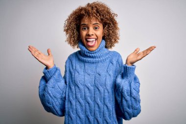 Beyaz arka plan üzerine balıkçı yaka kazak giyen genç ve güzel Afrikalı Amerikalı kadın çılgın ve hayret verici başarısını kollarını kaldırarak ve gözleri açık bir şekilde heyecanla haykırarak kutluyor. Kazanan konsept