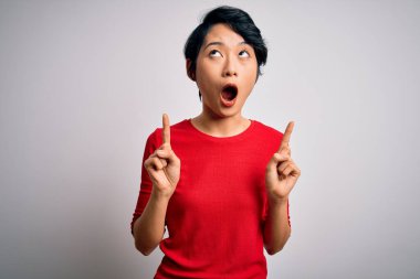 İzole edilmiş beyaz arka plan üzerinde duran gündelik kırmızı tişört giyen genç, güzel Asyalı kız şaşırmış ve şaşırmış bir şekilde yukarı bakıp parmaklarıyla işaret ediyor ve kollarını kaldırıyordu..