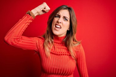 Genç ve güzel bir esmer kadın, kırmızı arka plan üzerine balıkçı yaka kazak giyiyor kızgın ve kızgın. Öfkeyle bağırırken yumruğunu kaldırıyor. Öfke ve agresif kavram.