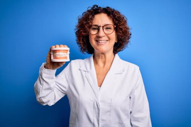 Mavi arka planda plastik diş takan orta yaşlı bir dişçi kadın. Mutlu bir yüzü var. Kendine güvenen bir gülümsemeyle dişlerini gösteriyor.