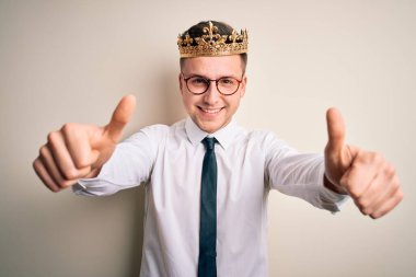 İzole edilmiş arka planda altın taç takan yakışıklı beyaz iş adamı el hareketleriyle olumlu jest yapmayı onaylıyor, gülümsüyor ve başarı için mutlu. Kazanan jest.