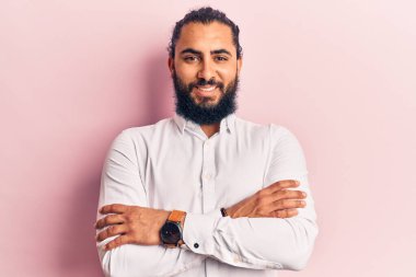 Genç Arap adam sıradan kıyafetler giyip kollarını kavuşturup kameraya bakarken gülümsüyor. pozitif kişi. 