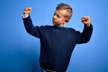 Mavi gözlü küçük beyaz çocuk mavi arka planda kışlık süveter giyiyor ve gururla gülümseyen kol kaslarını gösteriyor. Fitness konsepti.