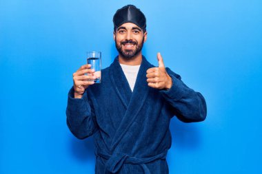 Uyku maskesi takan ve bornoz giyen genç adam gülümseyen ve pozitif gülümseyen, başparmağını kaldıran mükemmel ve onay işareti yapan. 