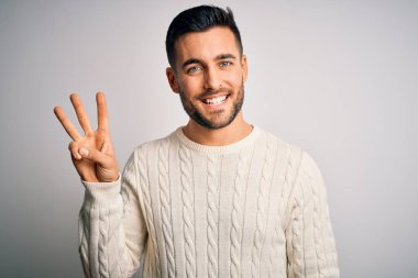 İzole edilmiş beyaz arka plan üzerinde duran sıradan süveter giyen genç ve yakışıklı bir adam üç numaralı parmaklarıyla işaret ederken kendinden emin ve mutlu gülümsüyor..